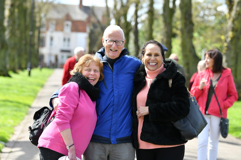 David is out on a walk with two other members. They have stopped to smile at the camera huddled in a group. A tree-lined path in a park is in the background.