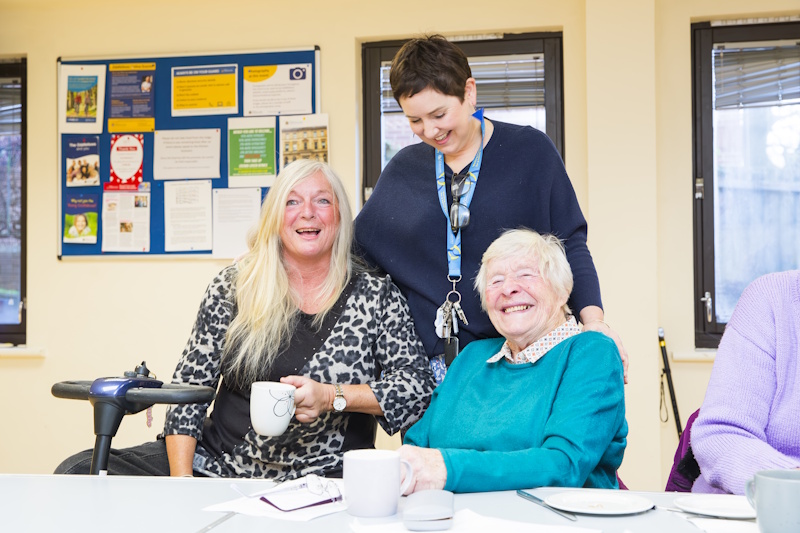 Three women laughing. One is stood between the other two are seated