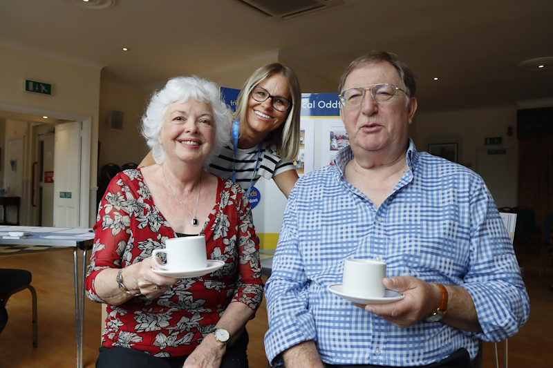 Two people sat down holding tea cups and one person stood behind them smiling