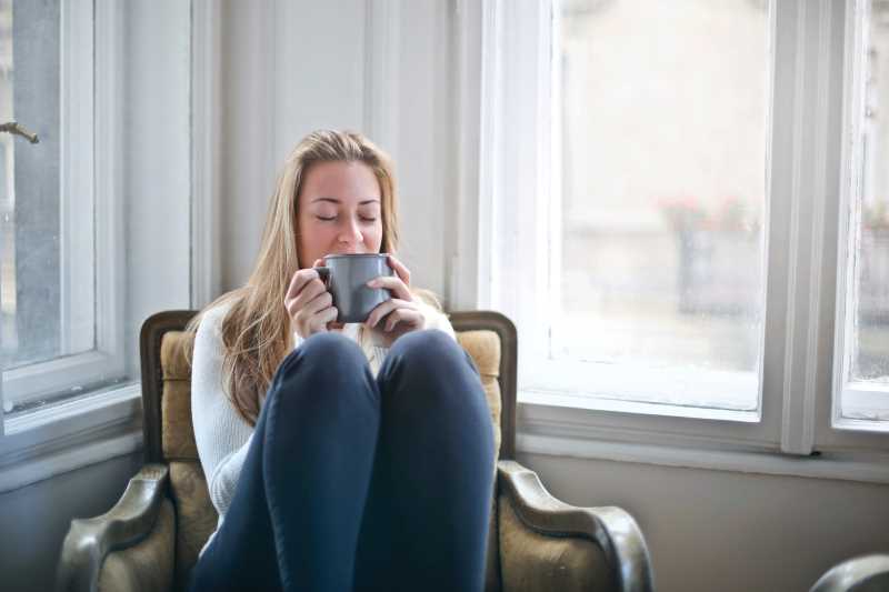 Woman in 30s sat knees up on an armchair winding down with a hot drink.