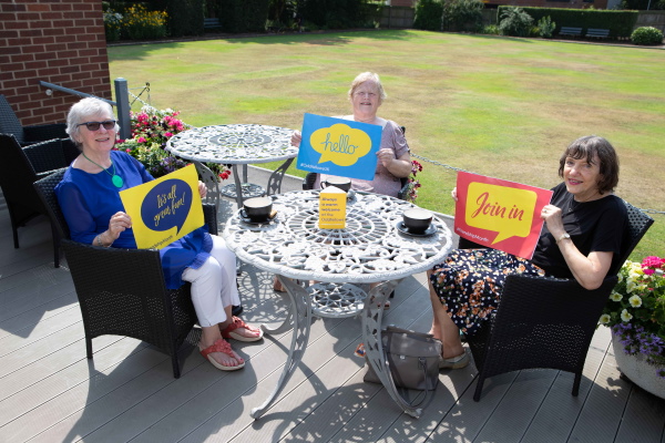 Three Oddfellows members sitting around a table outside and holding up signs which say "hello"