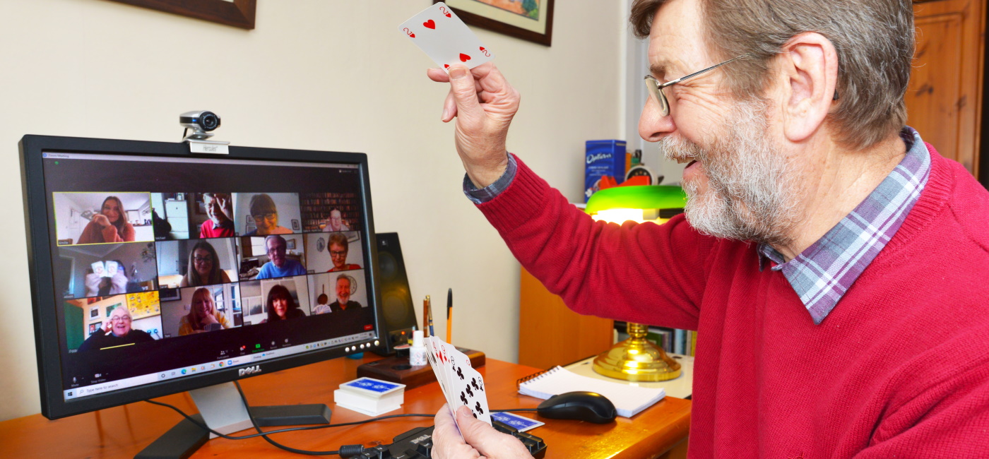 Man in 70s taking part in online zoom card event in front of a PC screen showing other players