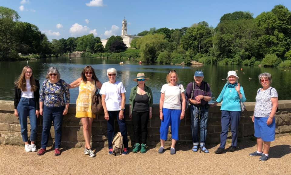 A line up of 9 members out on a walk in a park on a sunny day - in front of a lake.