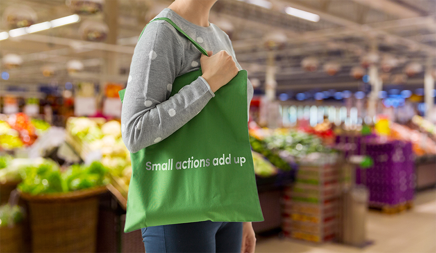 Woman shopping in a market holding a fabric tote bag which says small actions add up on it. Fruit and veg stalls in the background