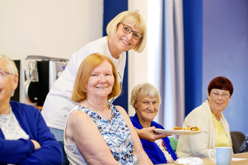 Vera is stood sharing a plate of cakes and biscuits with her friends who are sat at a table. All are smiling.