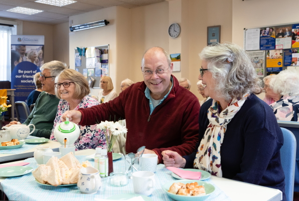 Kevin and Teresa at a branch afternoon tea event. Sat at  a table with other members and Kevin's pouring a cup of tea.