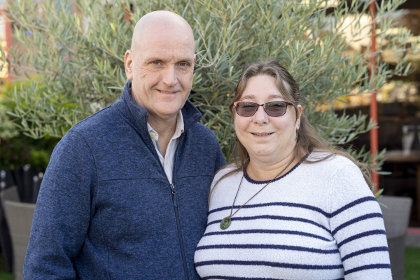 Jonathan and Dawn Platt stood together outside smiling with an olive tree behind them.