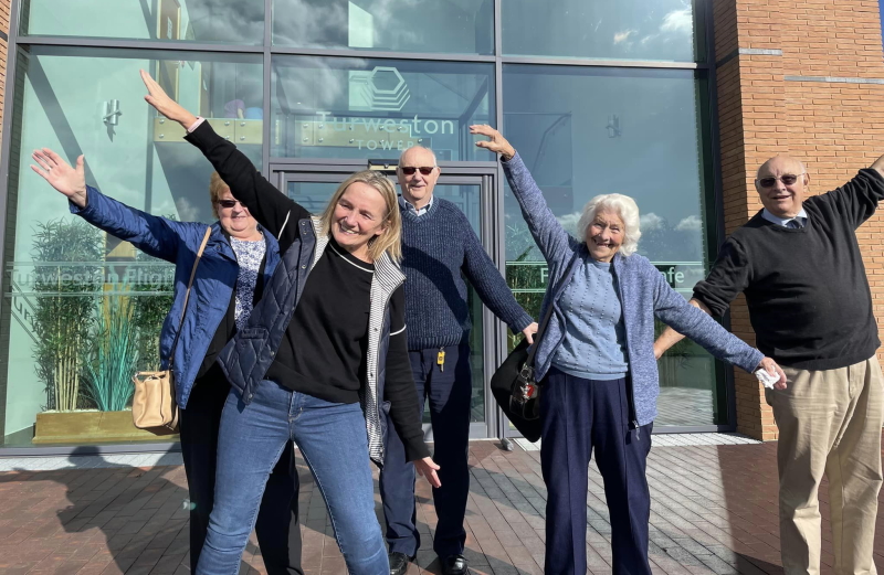 Five members stood in a group with their arms out like a plane flying, smiling