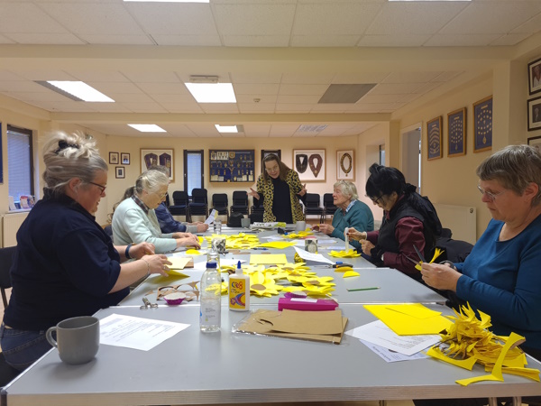 Rosemary stood up at the end of a large table while other women are seated, crafting paper sunflowers