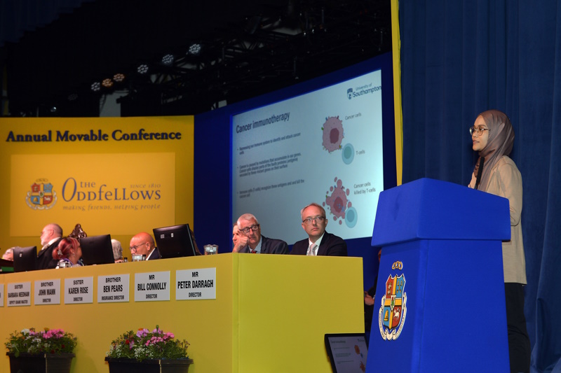 Nur stands behind a lectern and addresses the conference. Watching her talk are members of the Board