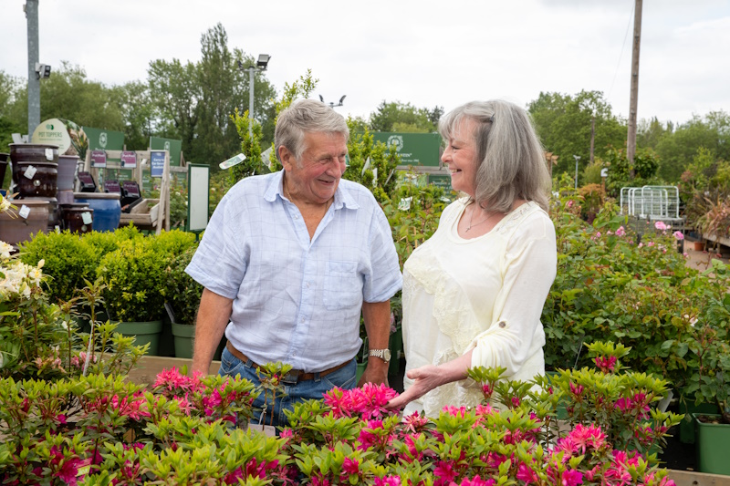 Ann stood with hisband, 'skip' next to some flowering shrubs at a garden centre