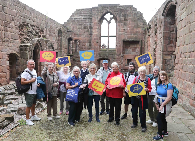 A group of people stood in some ruins smiling with Friendship Month placards