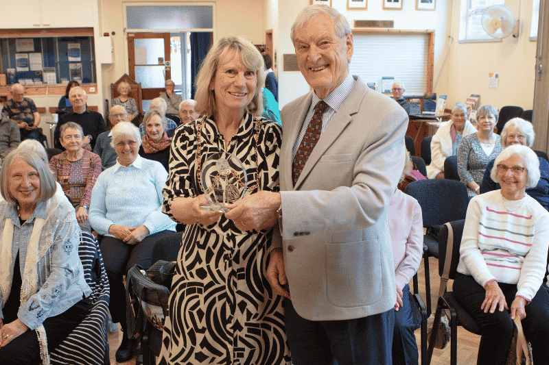 A man and a woman holding on to a glass trophy stood in front a group of people, all looking towards the camera