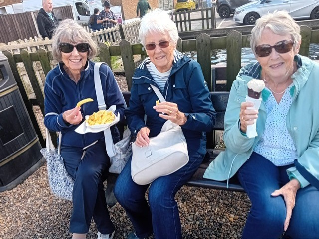 Three women at on a bench, one eating chips and another eating an ice cream