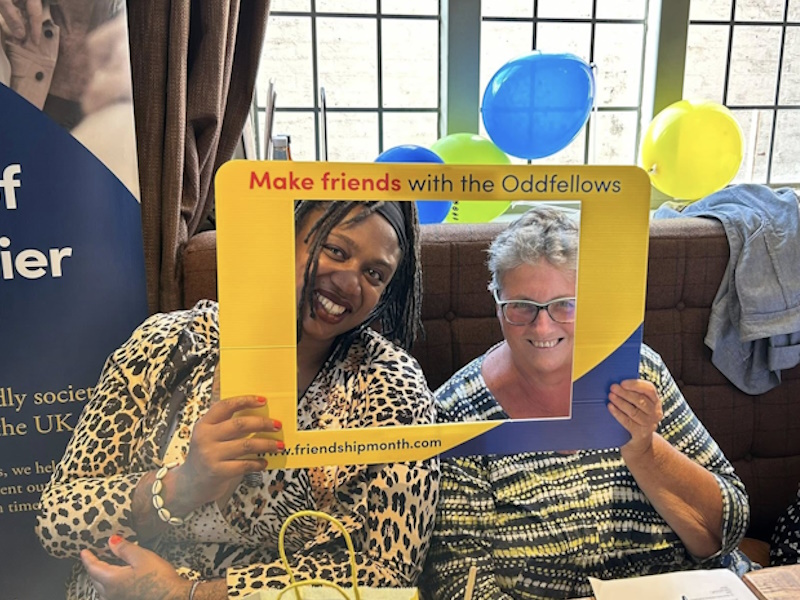 Two women smiling with a large frame around their faces which says 'make friends with the Oddfellows'