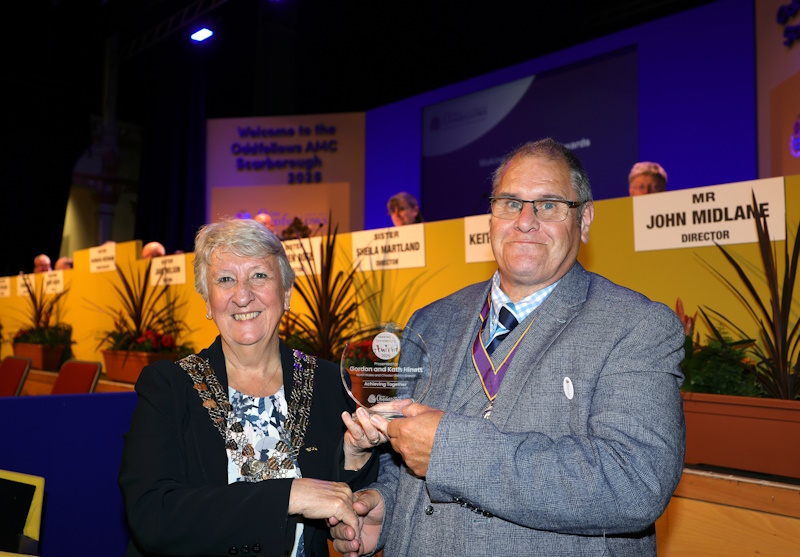A woman and man shaking hands both holding a trophy