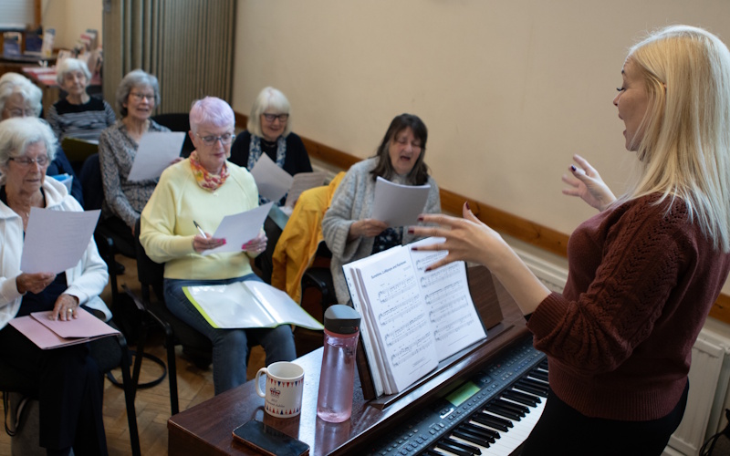 A woman stood by a piano, orchestrating a group of singers