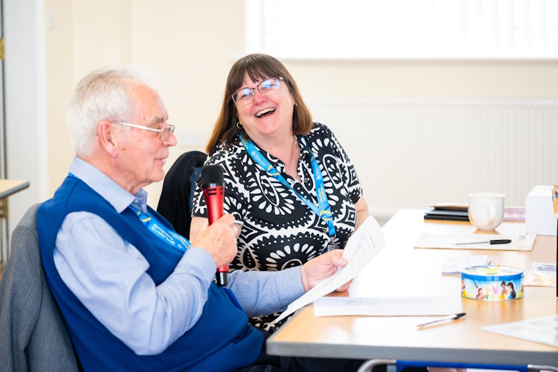 A man speaking into a microphone and a woman looking at him laughing