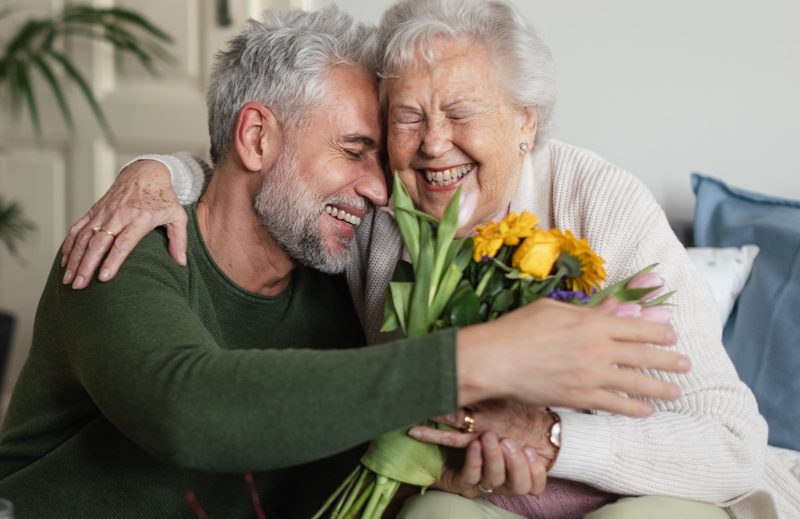 Man giving his older mum a hug and giving her a bunch of yellow flowers. Both are smiling and happy