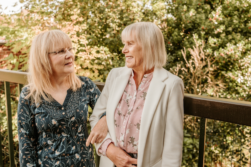 Jacqui and Eileen stood together linking arms and smiling at eash other