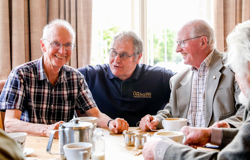 Gordon crouched down between two smiling men who are sat having coffee