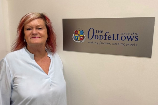 CEO of the Oddfellows, Jane Nelson, is stood next to a sign on the wall which displays the Oddfellows logo